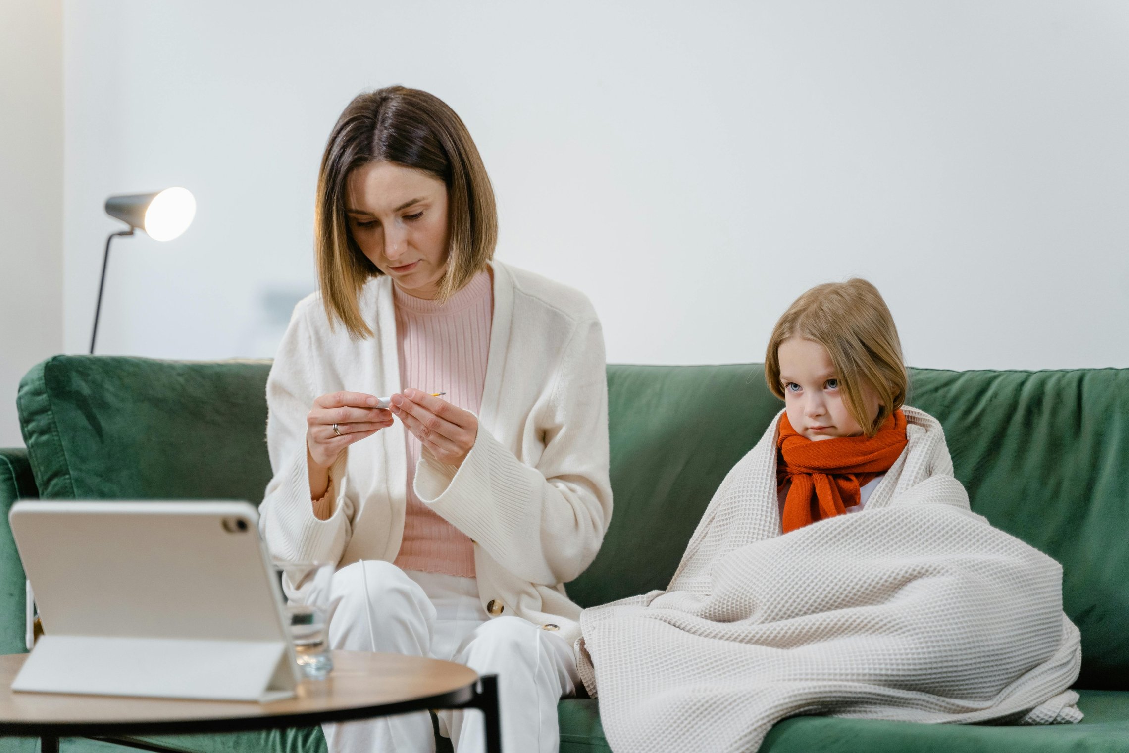 Moteris sėdi ant sofos ir žiūri į termometrą, šalia paklode apsisiautęs vaikas