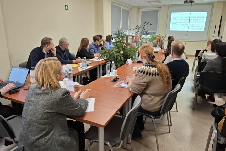 Ilgas stalas ir už jo sėdi žmonės su kompiuteriais ir rašymo priemonėmis